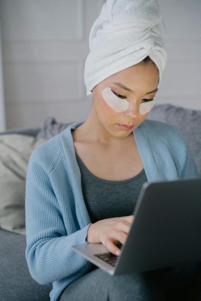 Woman applying skincare under-eye masks while using a laptop indoors.