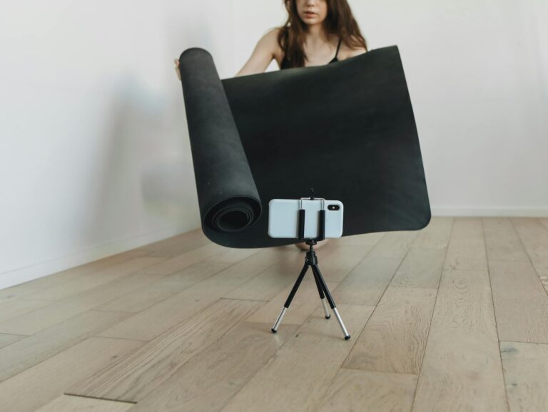 Woman rolls out yoga mat in living room with smartphone tripod for online session.